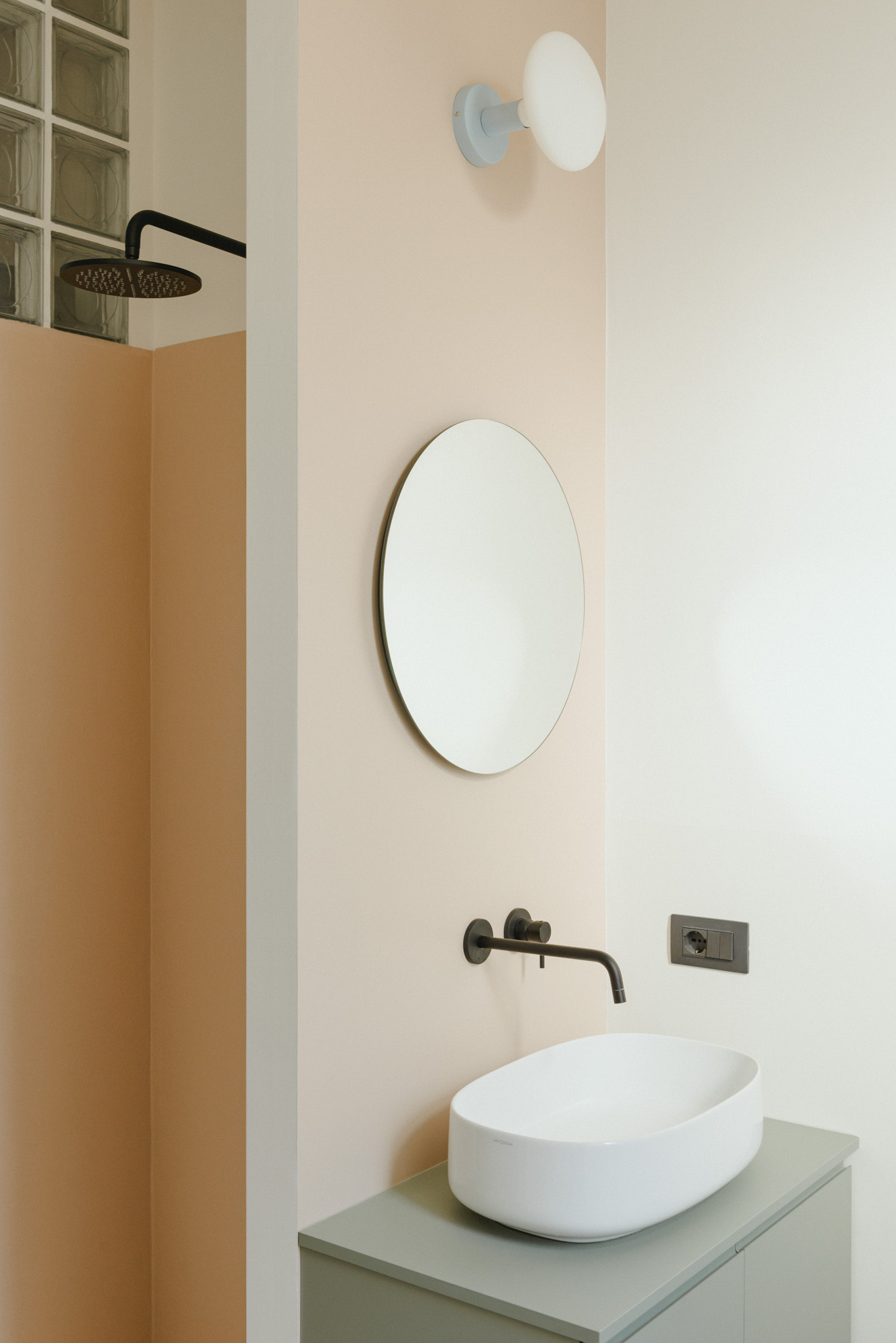 Modern cream bathroom with black faucet and shower, oval sink, circular mirror and sage green cabinet.