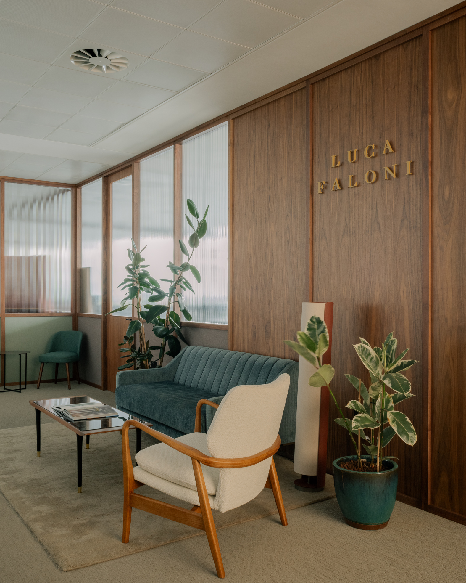 Entrance to an elegant office with a wooden armchair and light fabric, a wooden coffee table, a blue sofa, plants, and walls made of Canaletto walnut panels with fluted glass.