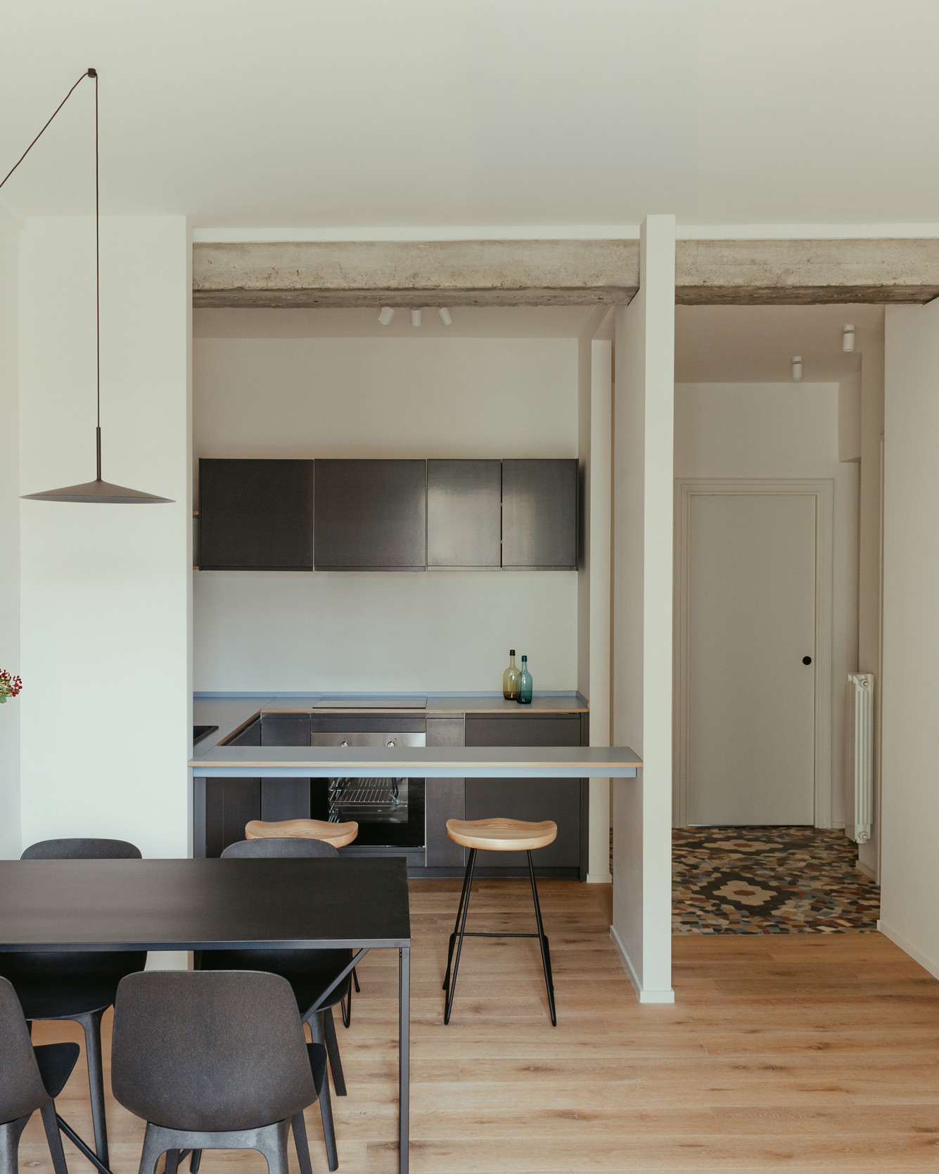 Minimalist dining room with light wood parquet, black table, light wood stool, suspended black lamp, and open black kitchen.