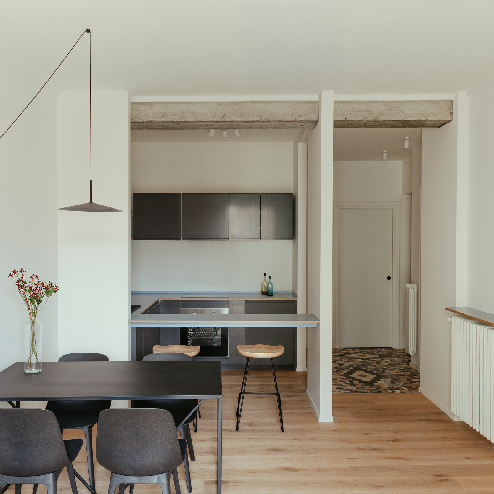 Minimalist dining room with light wood parquet, black table, light wood stool, suspended black lamp, and open black kitchen.