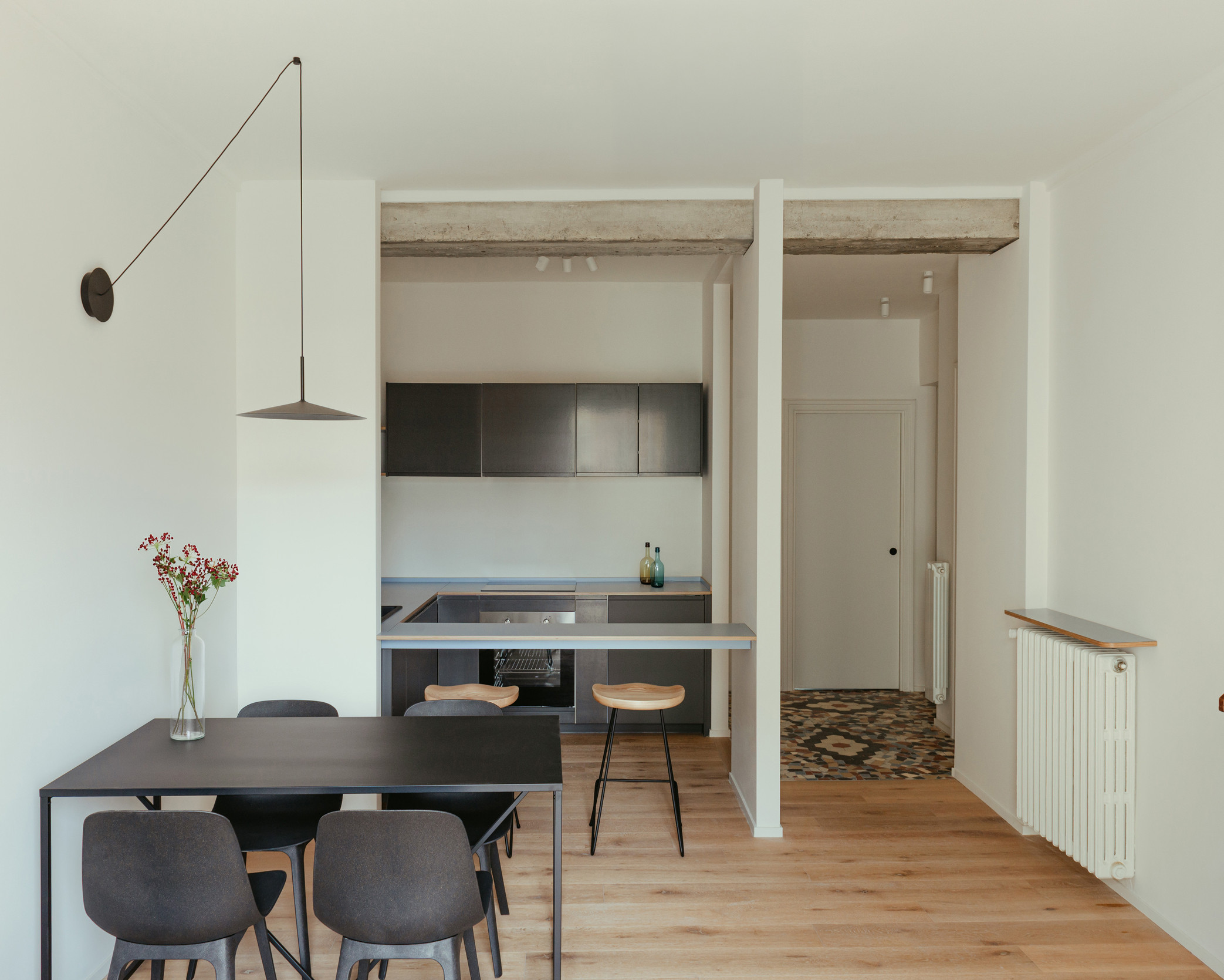 Minimalist dining room with light wood parquet, black table, light wood stool, suspended black lamp, and open black kitchen.