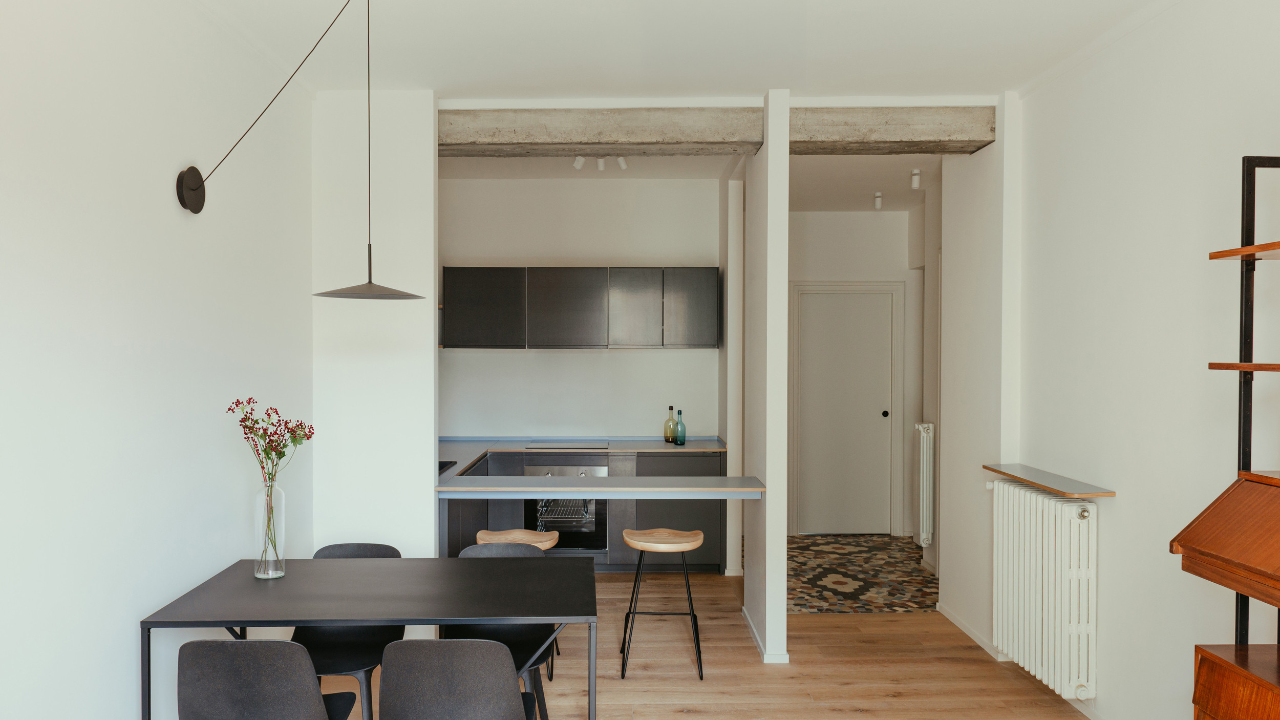 Minimalist dining room with light wood parquet, black table, light wood stool, suspended black lamp, and open black kitchen.
