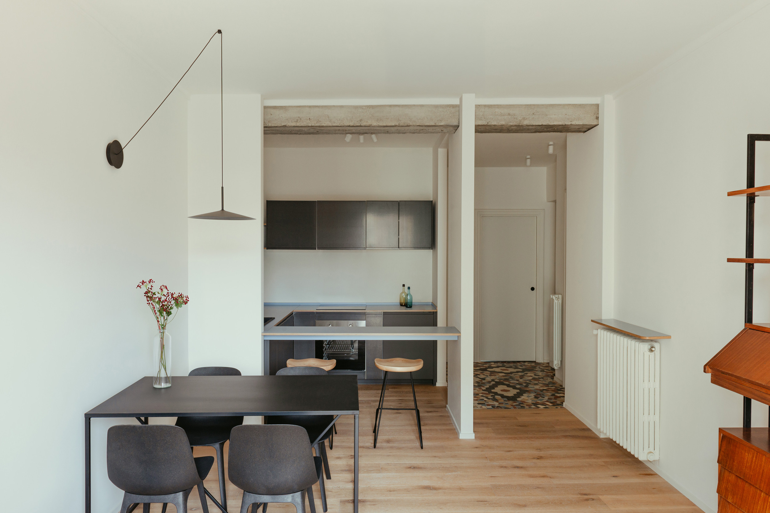 Minimalist dining room with light wood parquet, black table, light wood stool, suspended black lamp, and open black kitchen.